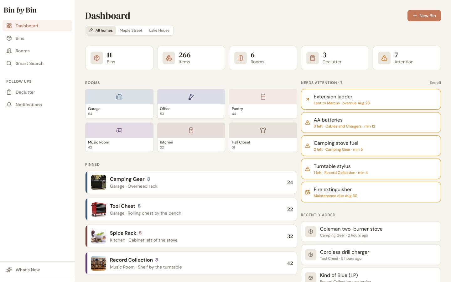 The Bin by Bin web dashboard on a laptop: a row of homes with All homes selected, counts for bins, items and rooms, a grid of rooms, pinned bins, and a Needs attention column listing an overdue loan and low-stock items.